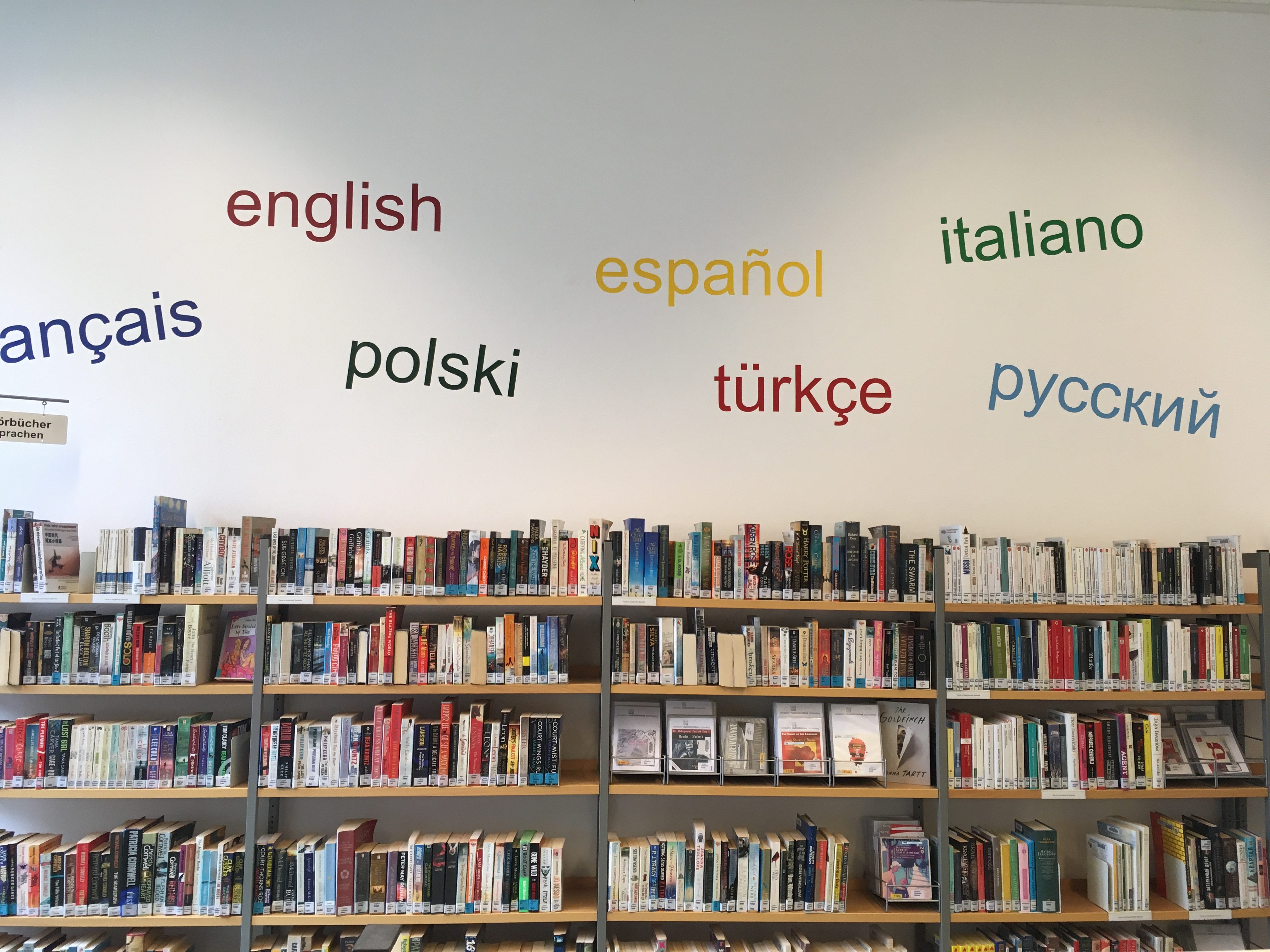 Shelves full of books in a library. The wall above has lettering reading "francais englisch polski espanol italiano..."