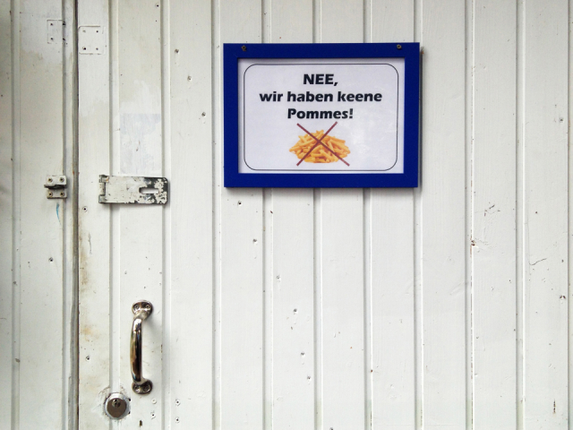 Sign on white wooden door reading "NEE, wir haben keene Pommes" with crossed-out picture of french fries